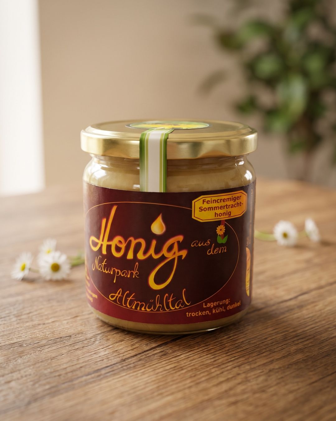 Ein Glas Feincremiger Sommertrachthonig mit dem Etikett „Honig aus dem Naturpark Altmühltal“ auf einem Holztisch, dahinter Gänseblümchen und eine Grünpflanze.