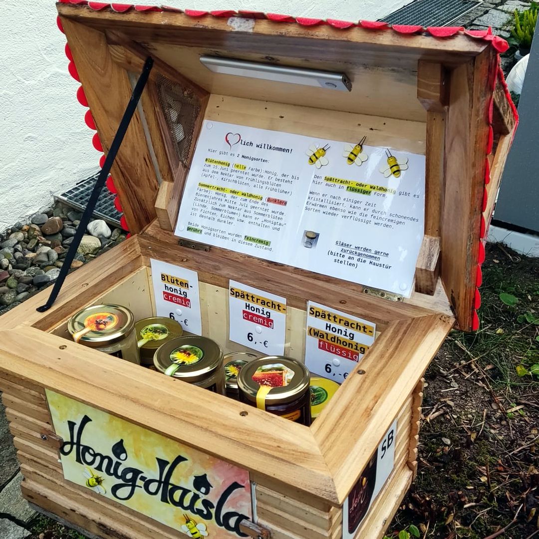 Das Honig-Haisla mit hochgeklapptem Dach: innen stehen Honiggläser vor Preisschildern mit 6 Euro, im Deckel hängt ein Begrüßungszettel.