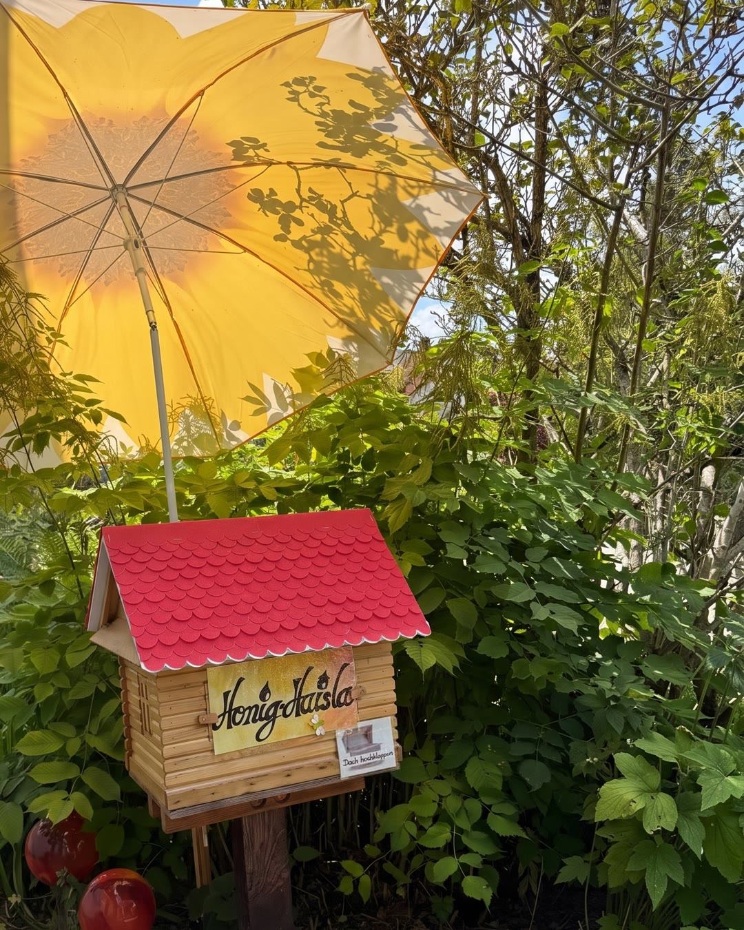 Das Honig-Haisla: ein kleines Holzhäuschen mit rotem Schindeldach auf einem Pfosten, darüber ein Sonnenschirm mit Sonnenblume, ringsum Grün.