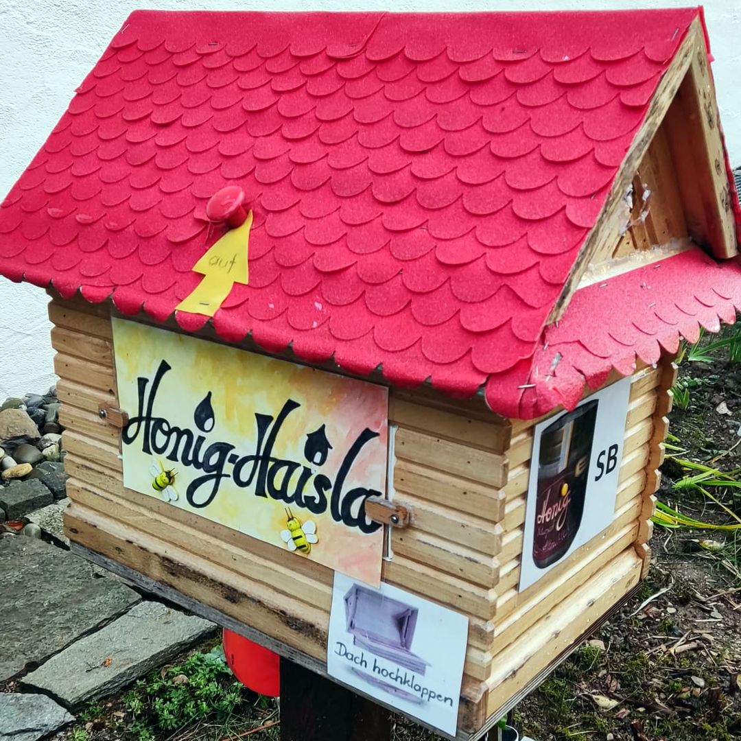 Das Honig-Haisla geschlossen: rotes Schindeldach mit einem gelben Pfeil „auf“ am Griff, vorn das Schild Honig-Haisla und der Zettel „Dach hochklappen“.